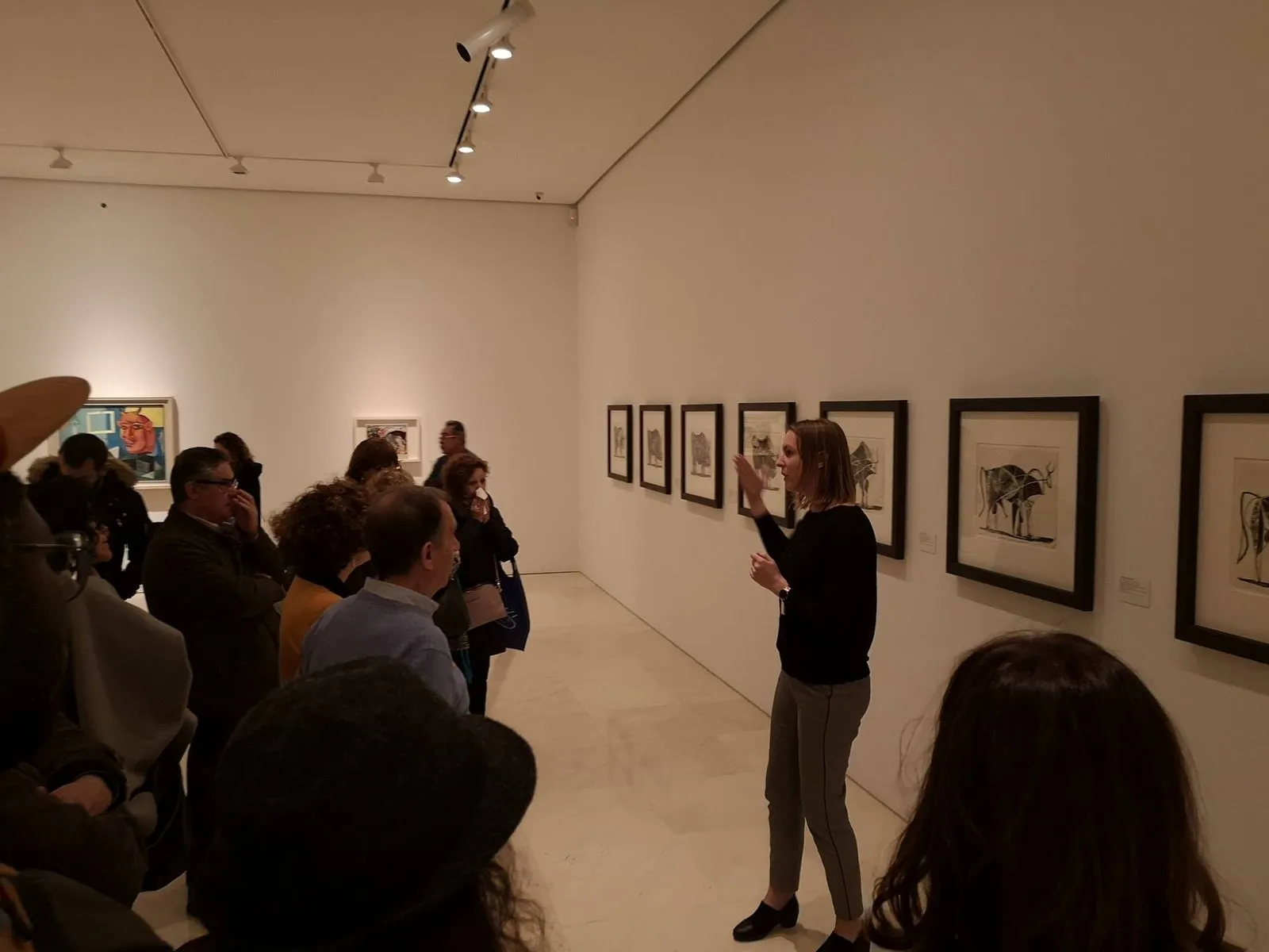 Anaïs raises a hand towards a line of framed etchings while the group follows along the gallery wall.