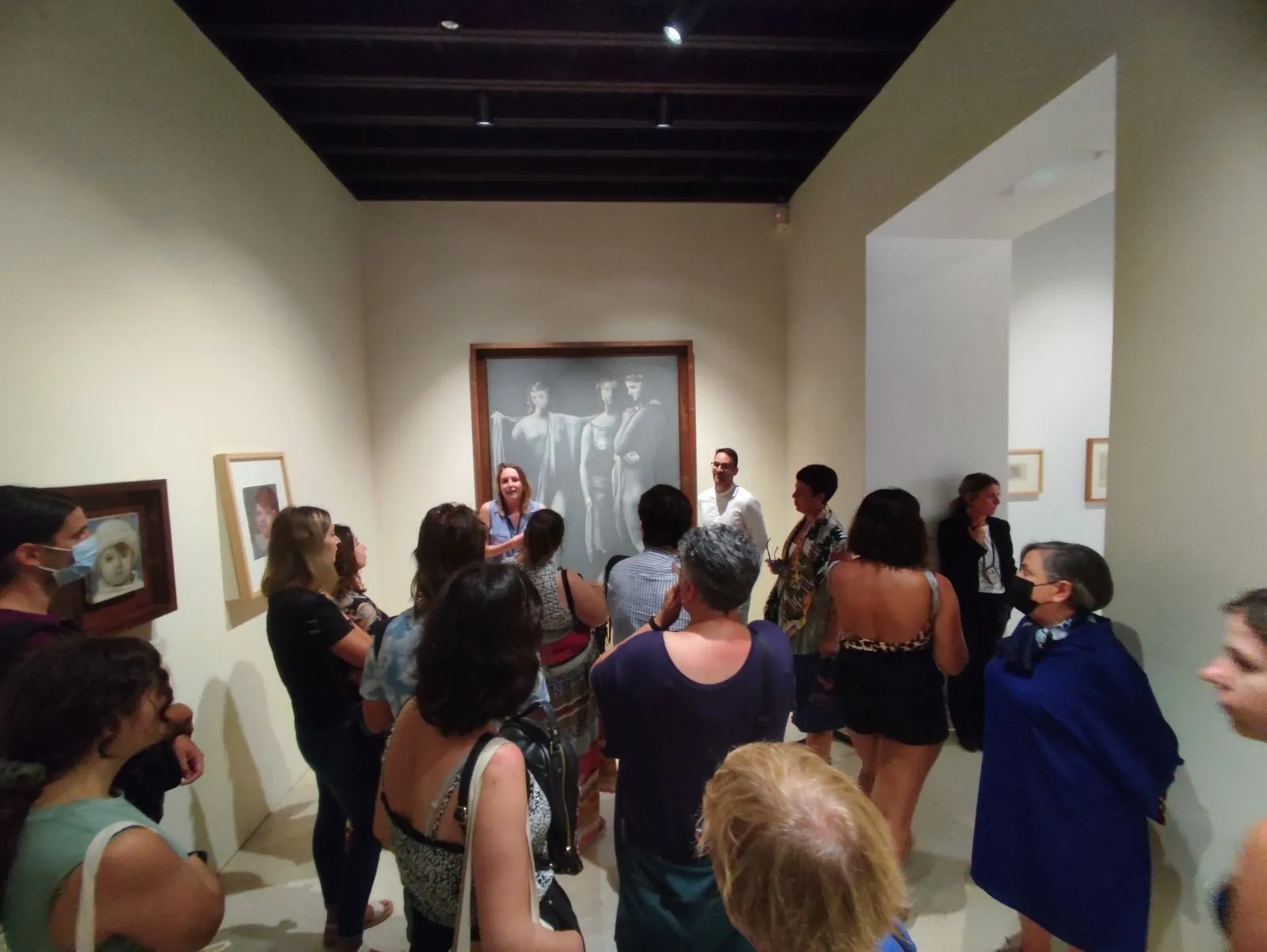 A room under dark wooden beams, a large grey canvas of three figures on the far wall behind the visitors.