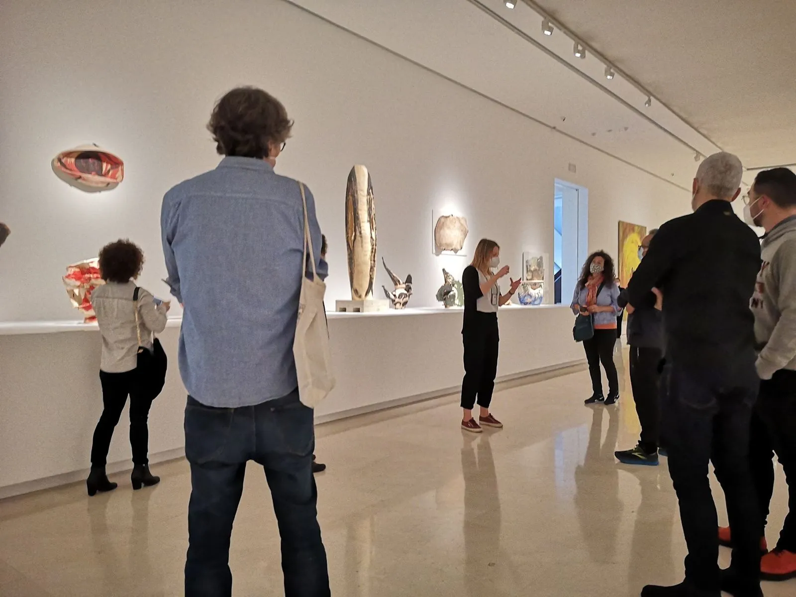 Ceramic pieces lined up along a long white ledge in a bright room, visitors listening a few steps back.