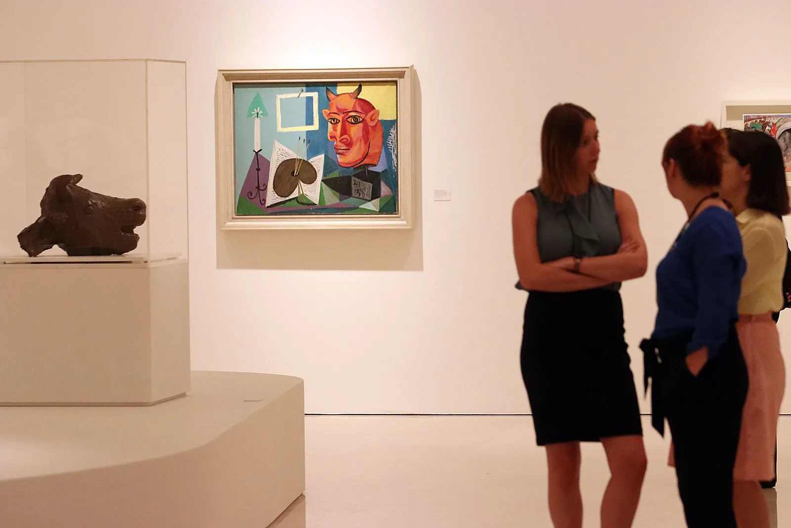 A bronze bull head in a vitrine beside a Picasso canvas of an orange horned face, three visitors talking further down the room.